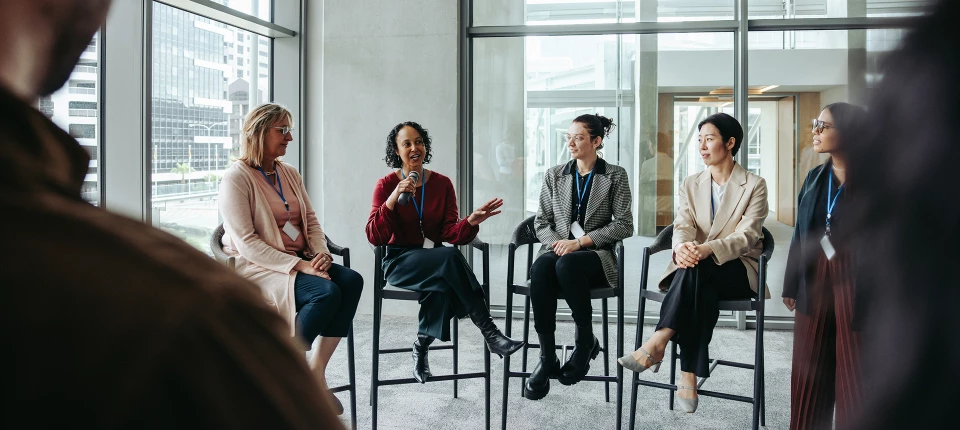 Deux femmes assises sur des tabourets haut en train de discuter devant d'autres personnes, la femme de gauche à la parole et parle dans un micro